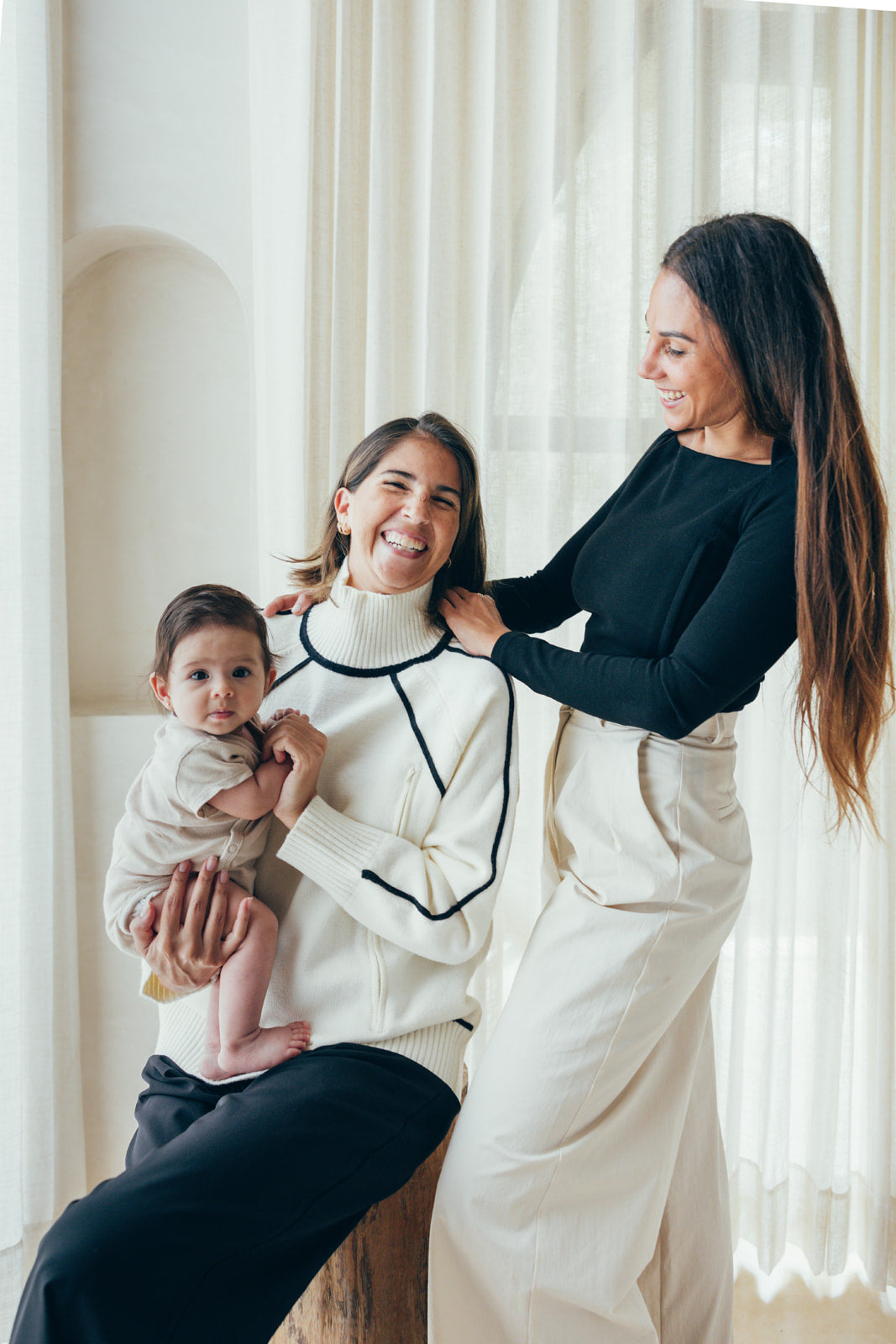Maman assise portant le pull d'allaitement blanc Valentine 23 Mai Paris avec son bébé dans les bras, accompagnée de sa copine debout à côté, riant ensemble.